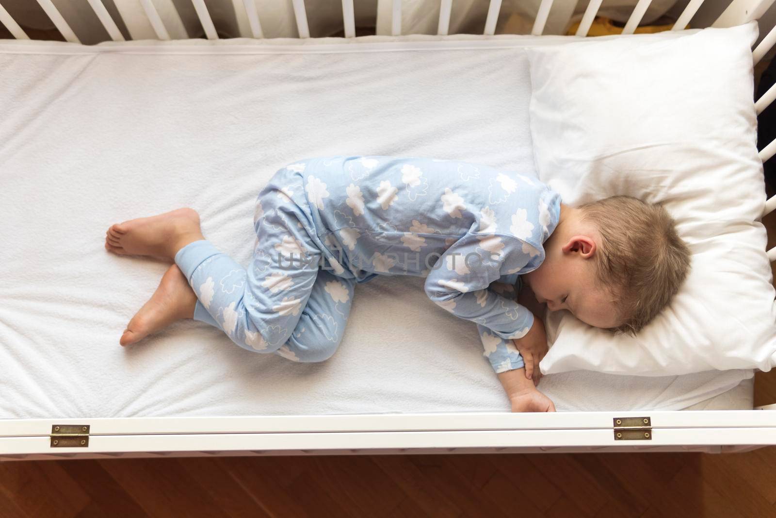 top view Cute little 2-3 years preschool baby boyl kid sleeping sweetly in white crib during lunch rest time in blue pajama with pillow at home. Childhood, leisure, comfort, medicine, health concept.