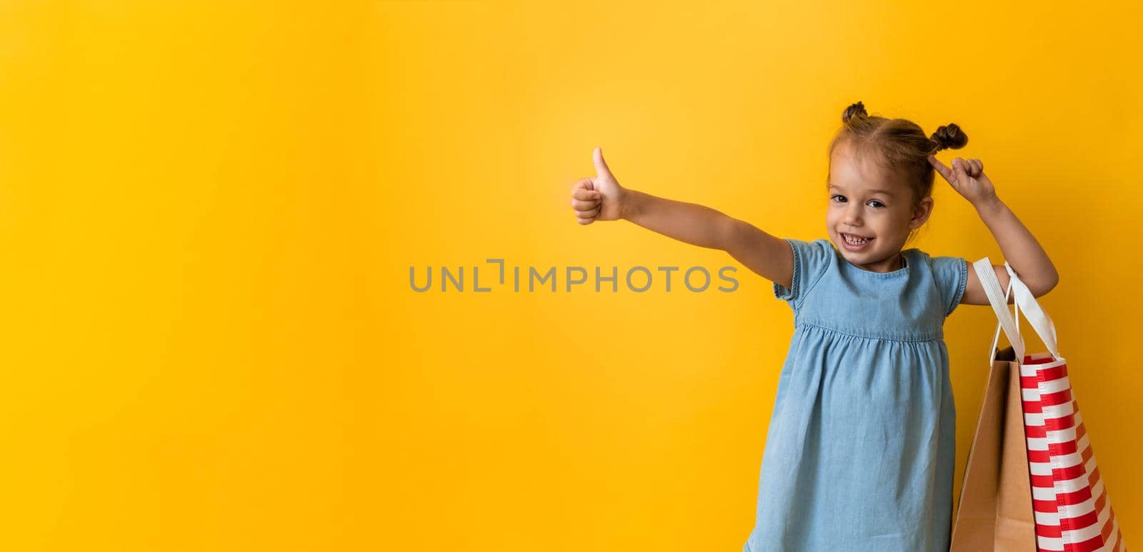 Banner Portrait Caucasian Beautiful Happy Little Preschool Girl Smiling Cheerful And Holding Cardboard Bags Thumb Up On Orange Yellow Background. Happiness, Consumerism, Sale People shopping Concept.