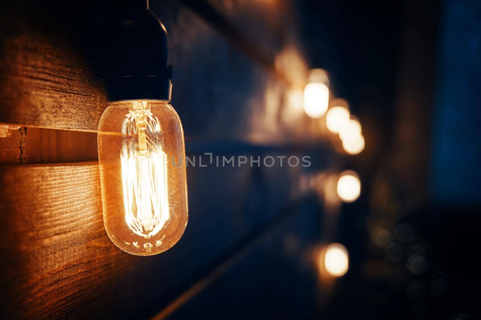 Antique incandescent Edison type bulb on wooden texture wall. Blurred warm garland light on background. Christmas night party mood. Holiday interior decorating. Copy space for congratulations.