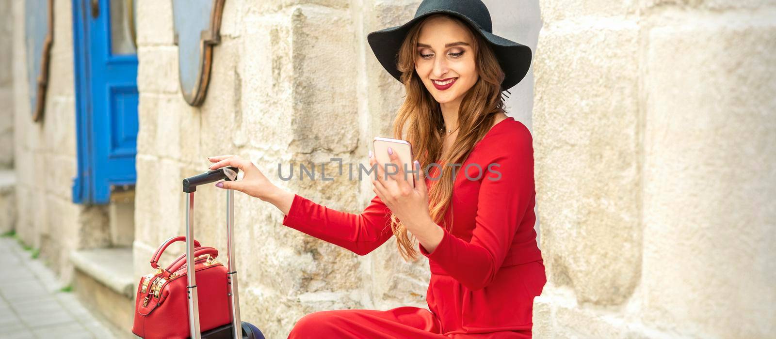 Beautiful young caucasian traveling woman in black hat looking on the smartphone smiling and sitting near the building on the street outdoors