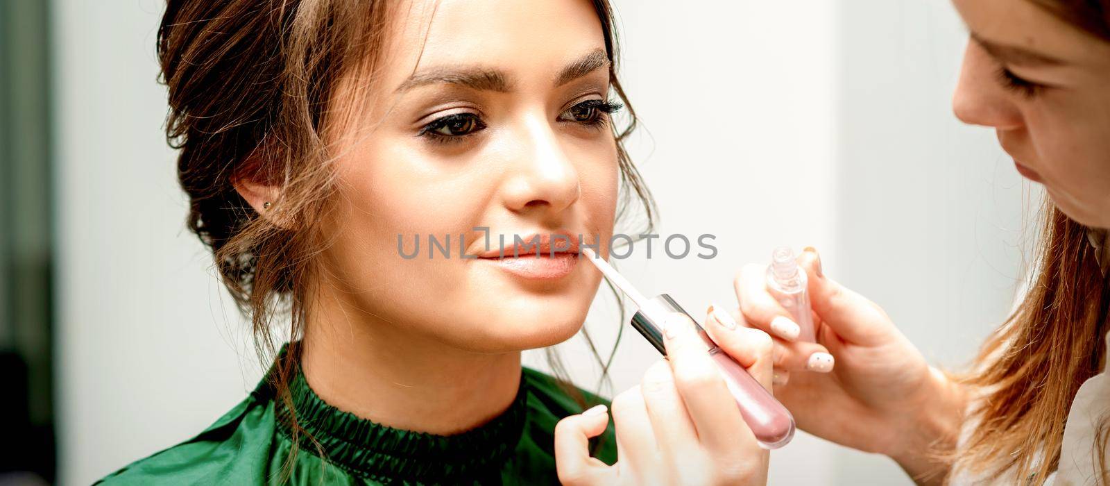 Close up of makeup artist applying lipstick foundation with a make-up brush on lips of the beautiful young caucasian woman in a beauty salon