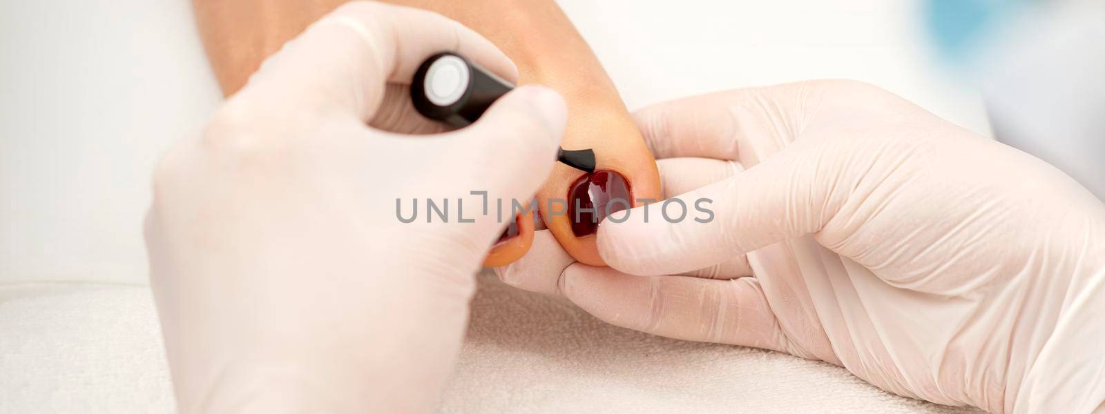 Closeup of hands in white gloves applying burgundy nail polish with a brush on woman's toes in a nail salon
