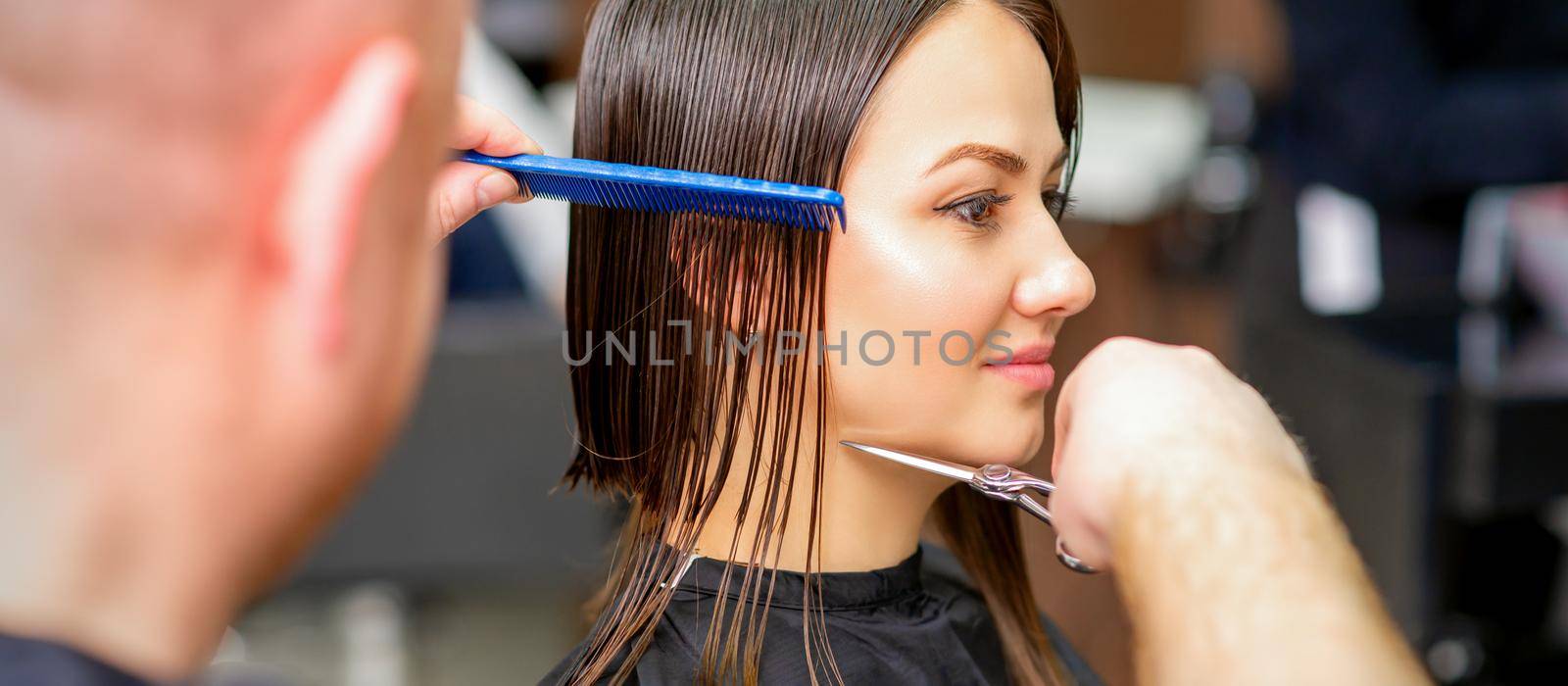 Male hairdresser cuts wet hair of young caucasian woman combing with a comb in a hair salon