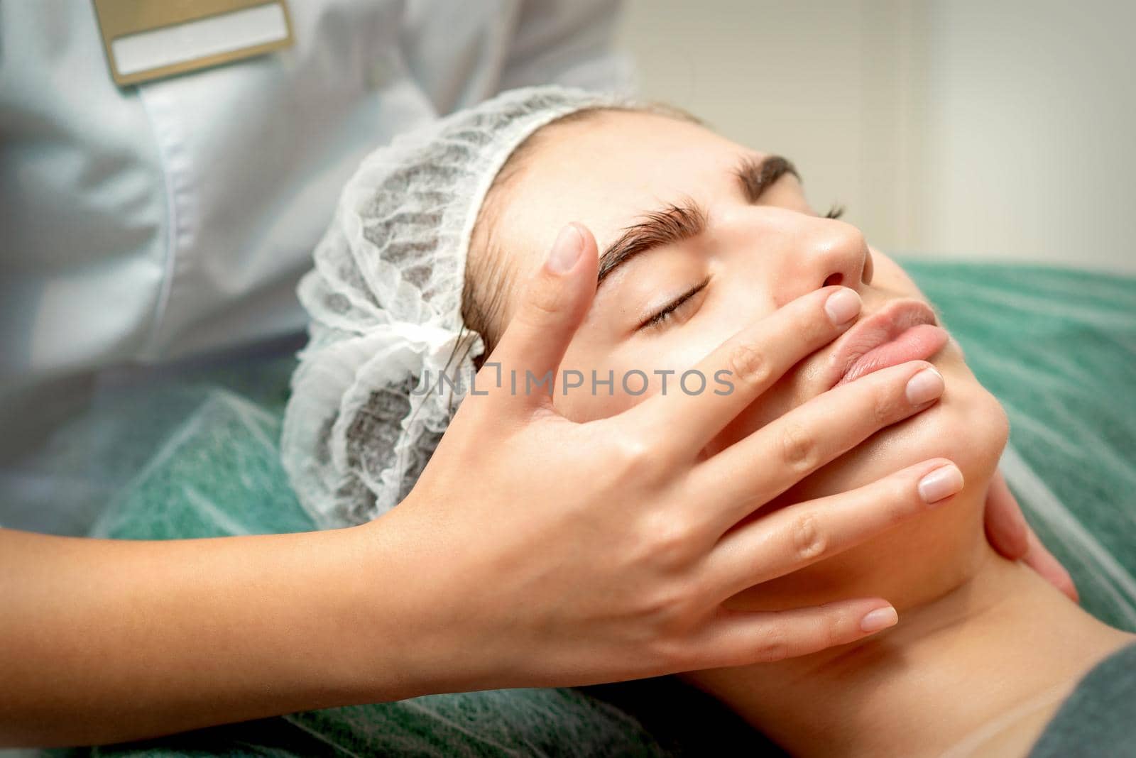 Young caucasian woman receiving facial massage by beautician's hands in spa medical salon