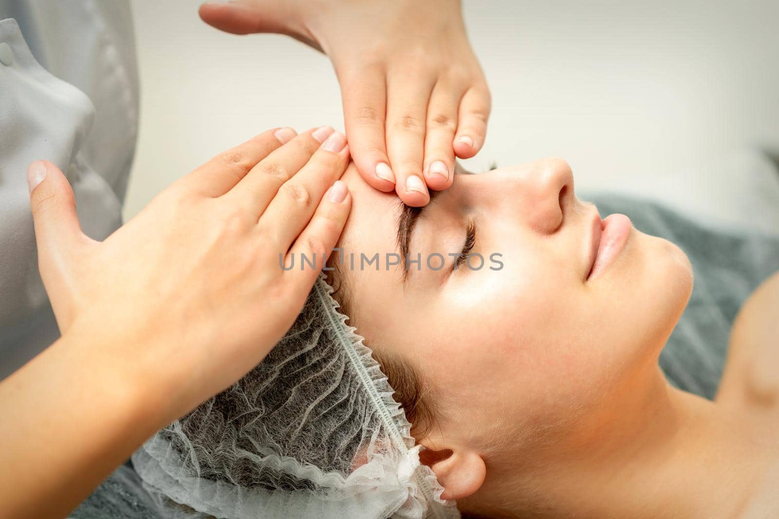 Young caucasian woman receiving facial massage by beautician's hands in spa medical salon