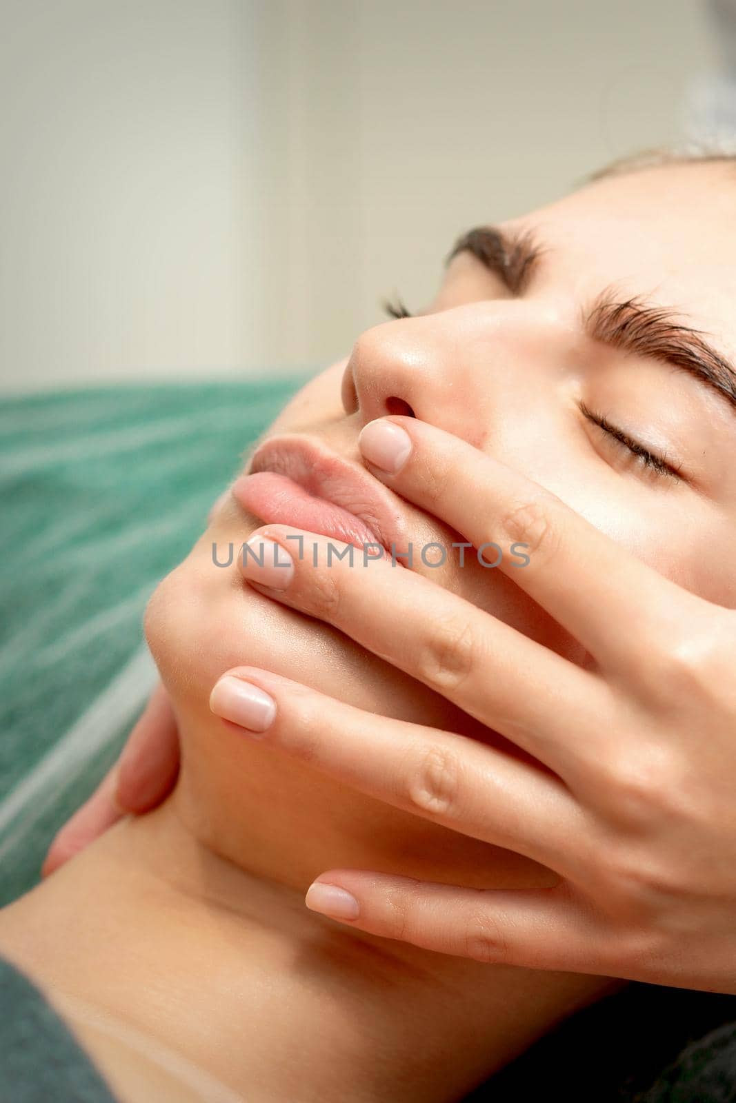 Young caucasian woman receiving facial massage by beautician's hands in spa medical salon