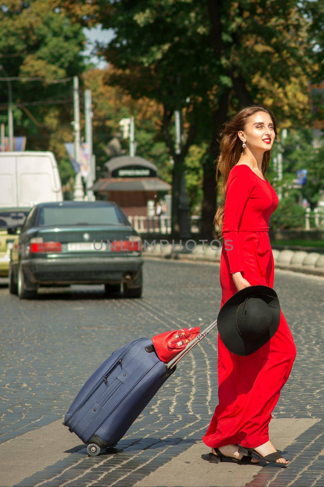 Beautiful young caucasian tourist woman with a suitcase in red long dress crosses the road through a crosswalk on the city street outdoors