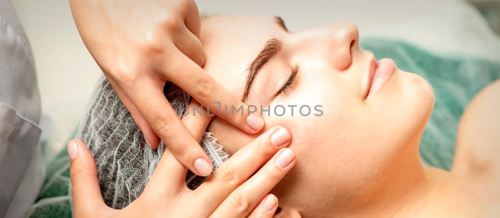 Young caucasian woman receiving facial massage by beautician's hands in spa medical salon