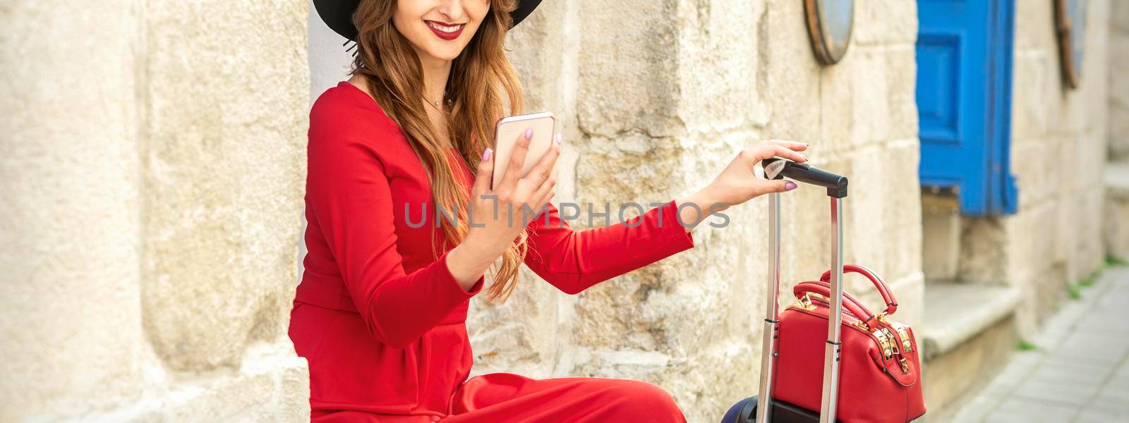 Beautiful young caucasian traveling woman in black hat looking on the smartphone smiling and sitting near the building on the street outdoors