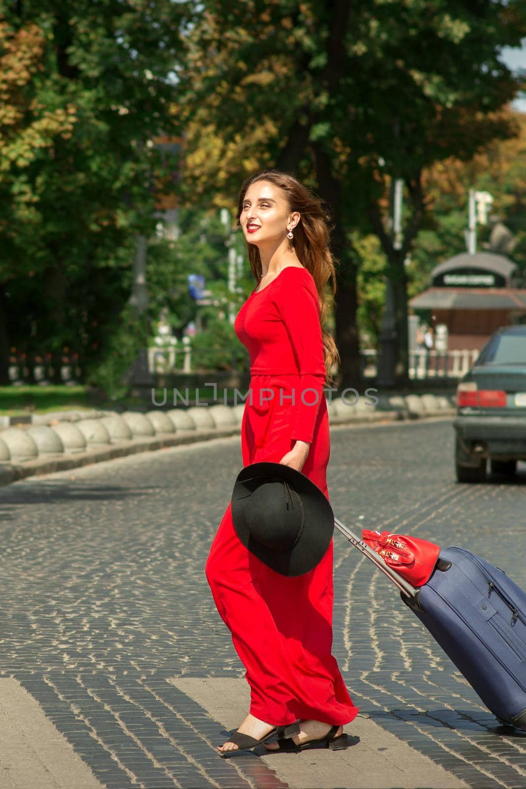 Beautiful young caucasian tourist woman with a suitcase in red long dress crosses the road through a crosswalk on the city street outdoors