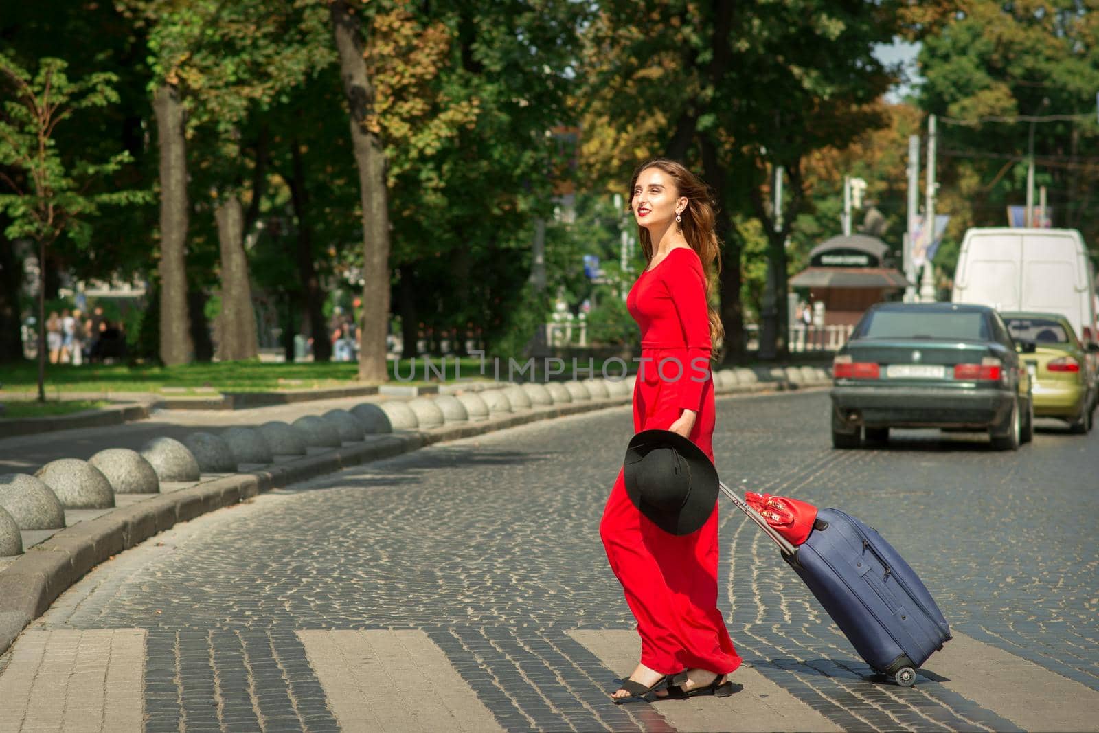 Beautiful young caucasian tourist woman with a suitcase in red long dress crosses the road through a crosswalk on the city street outdoors