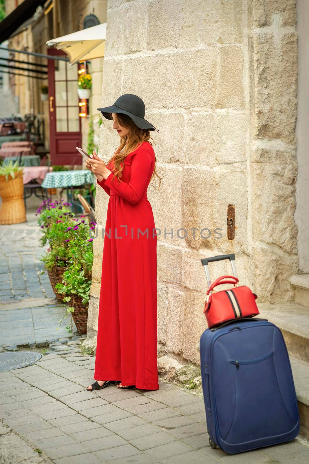 Beautiful young caucasian travel woman in a red long dress with suitcase standing and looking in a smartphone on the old city street