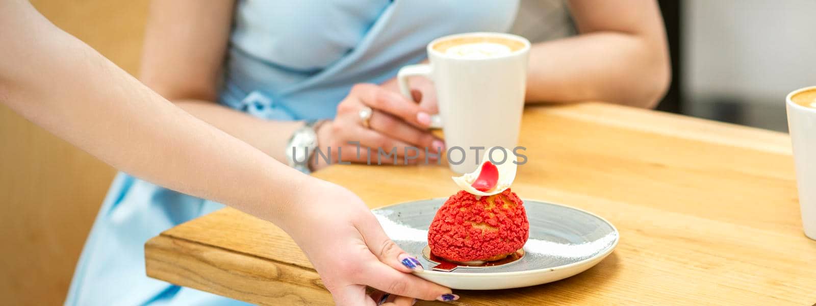 The waiter's hand puts on the table a plate with a red cupcake on the background of a female client in a cafe