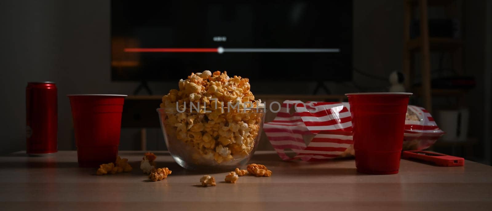 Bowl of tasty popcorn and cups on wooden table in living room. Family vacation, recreation, relaxation and hobby concept.