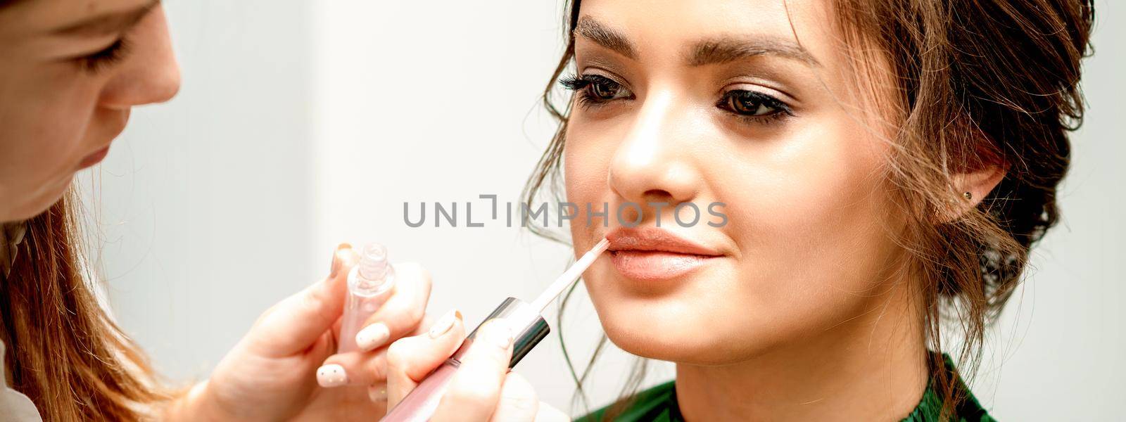 Close up of makeup artist applying lipstick foundation with a make-up brush on lips of the beautiful young caucasian woman in a beauty salon