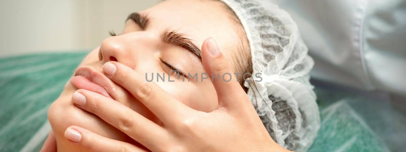 Young caucasian woman receiving facial massage by beautician's hands in spa medical salon