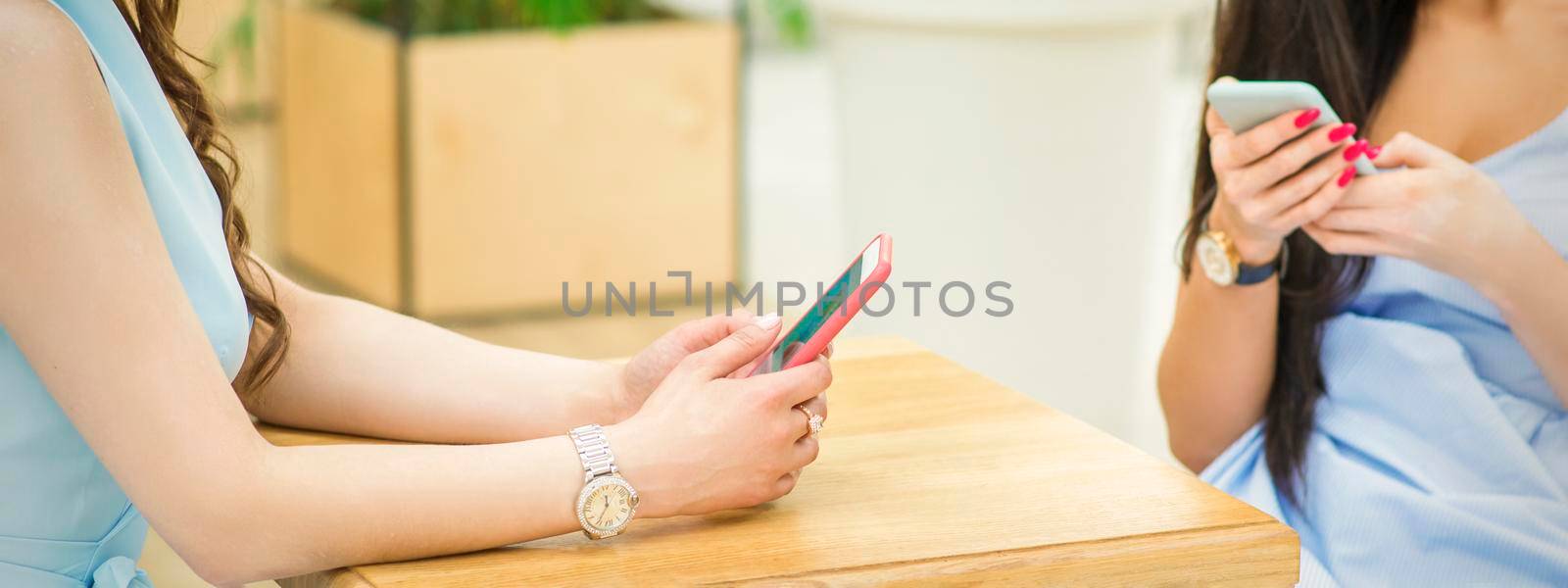 Hands of two young white women holding smartphones sitting at the table outdoors