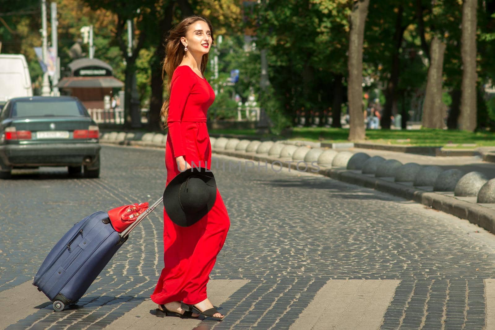 Beautiful young caucasian tourist woman with a suitcase in red long dress crosses the road through a crosswalk on the city street outdoors