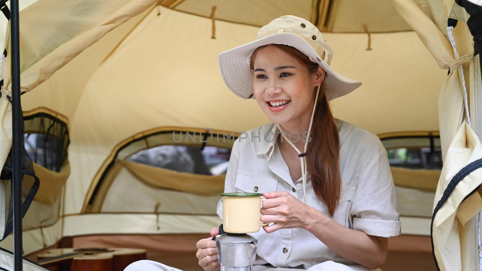 Pleasant asian woman sitting inside camp tent and drinking coffee. Travel, adventure and vacation concept by prathanchorruangsak