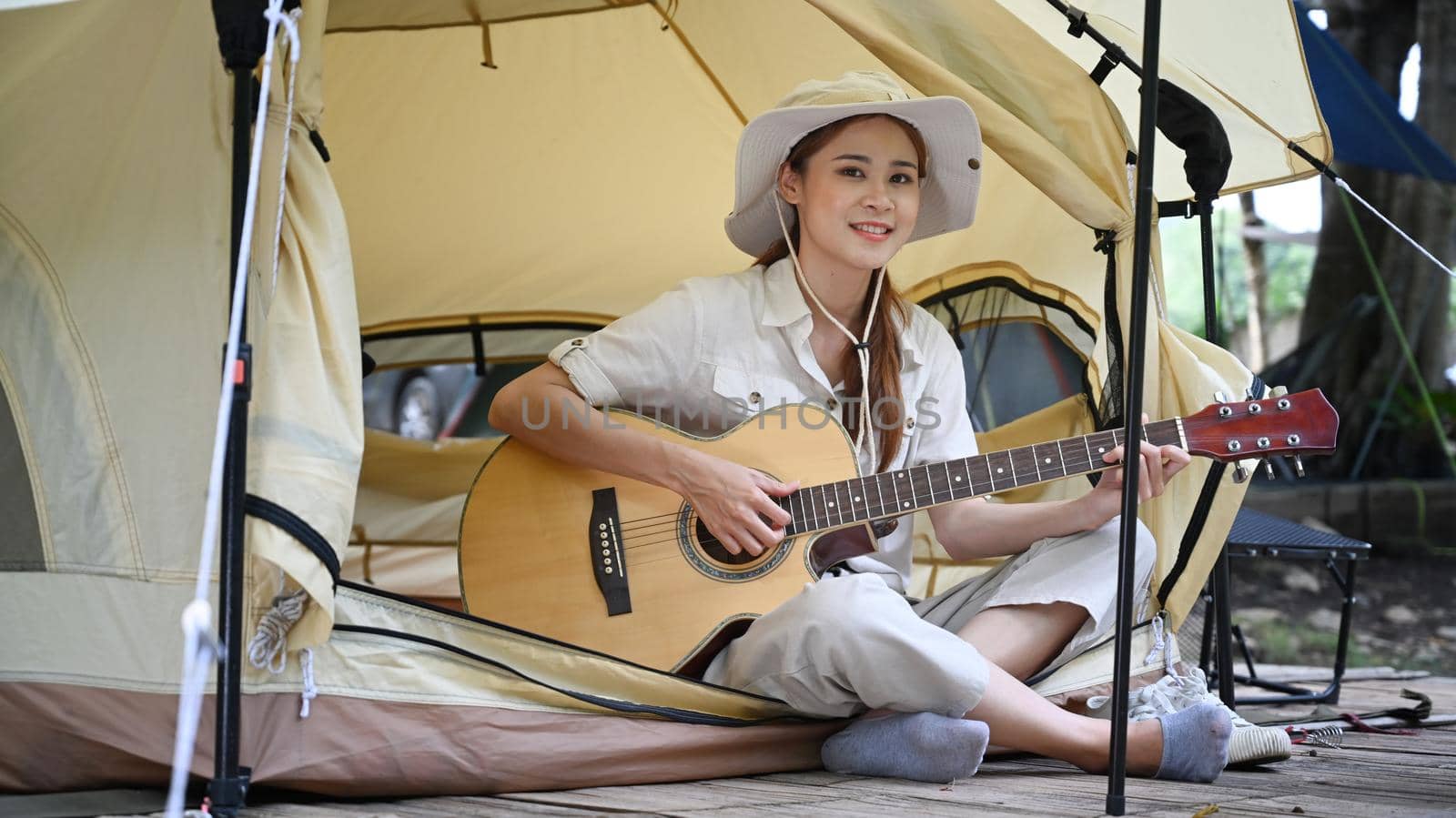 Happy young woman playing guitar while sitting inside camping tent. Travel, adventure and vacation concept by prathanchorruangsak