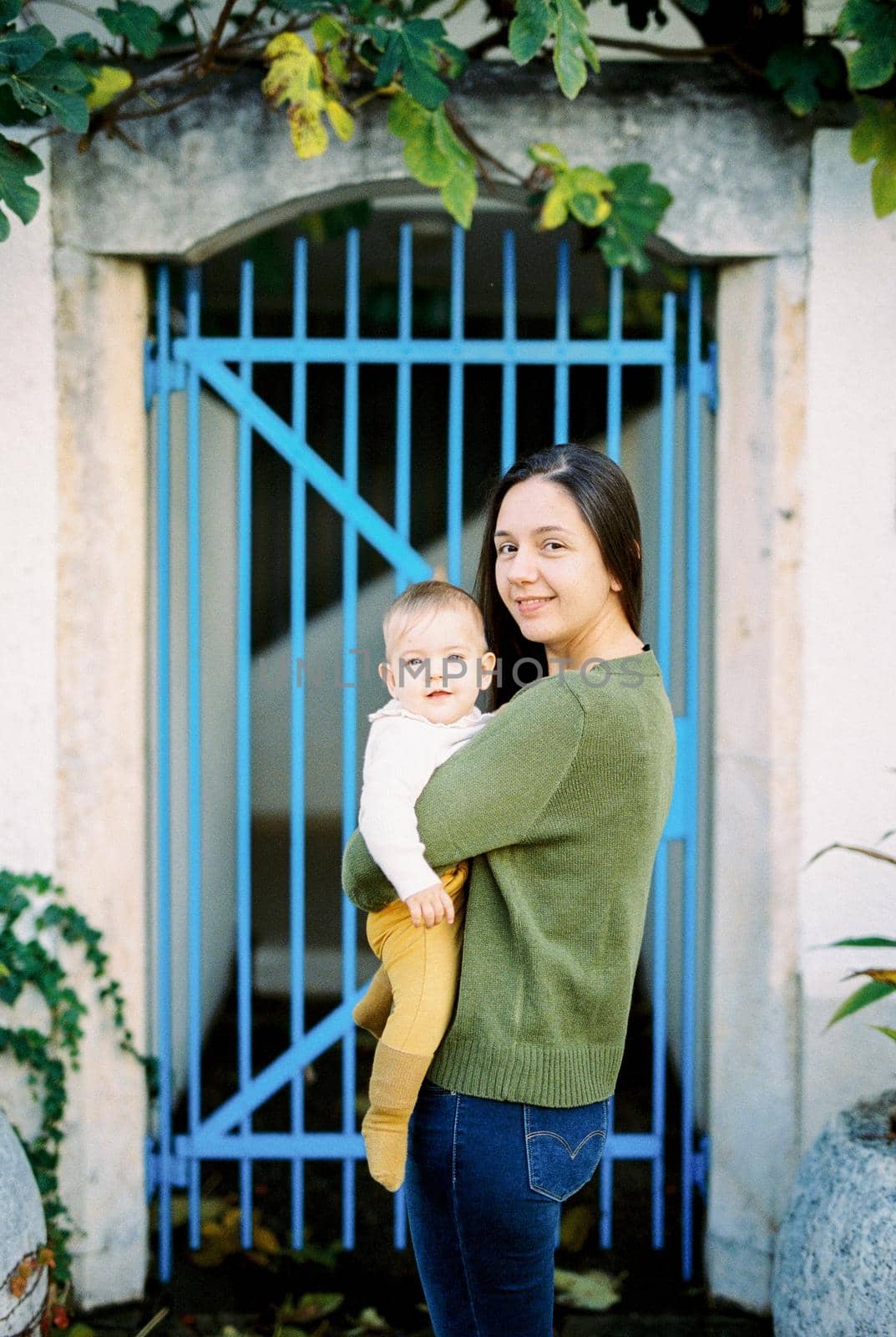 Mom with a baby in her arms stands near a forged gate by Nadtochiy