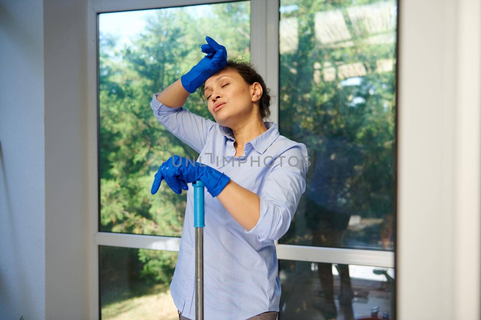 Stunning attractive multi-ethnic woman, lovely housewife, maid in rubber work gloves feeling exhausted, tired after house cleaning. Household. Housekeeping. Services of professional cleaning company