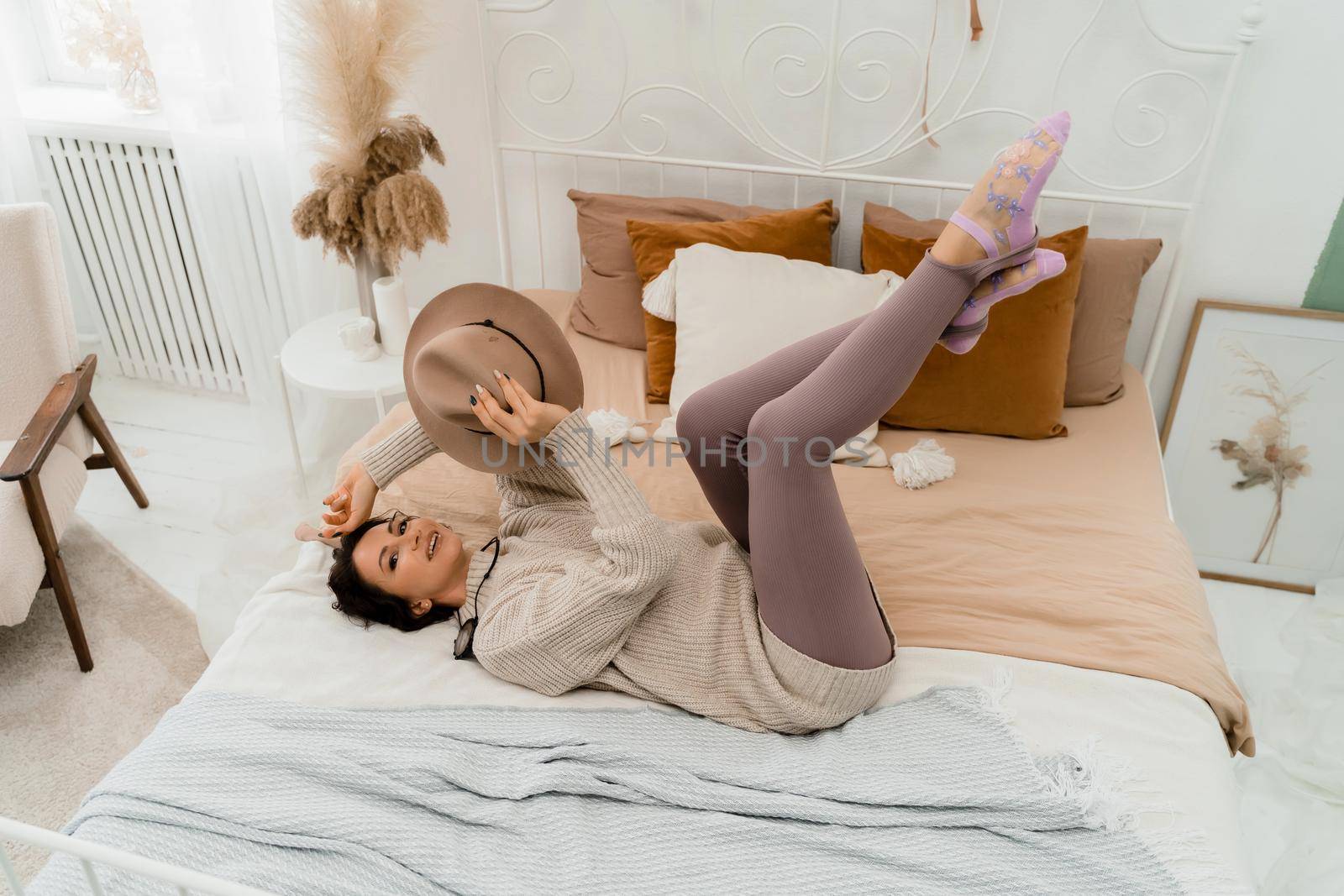 Lifestyle concept. A young middle-aged woman in a sweater lies on the bed and laughs, holds a brown hat in her hands