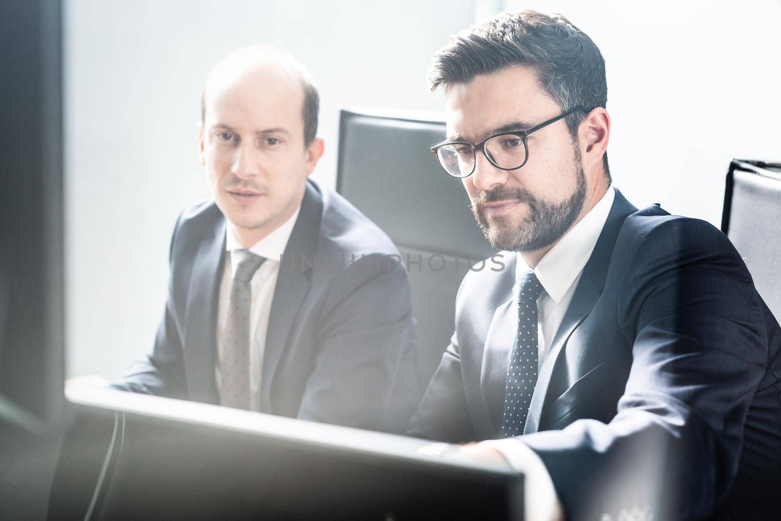 Image of two thoughtful businessmen looking at data on multiple computer screens, solving business issue at business meeting in modern corporate office. Business success concept.