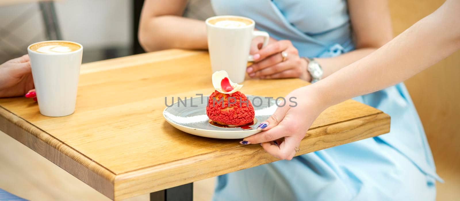 The waiter's hand puts on the table a plate with a red cupcake on the background of a female client in a cafe
