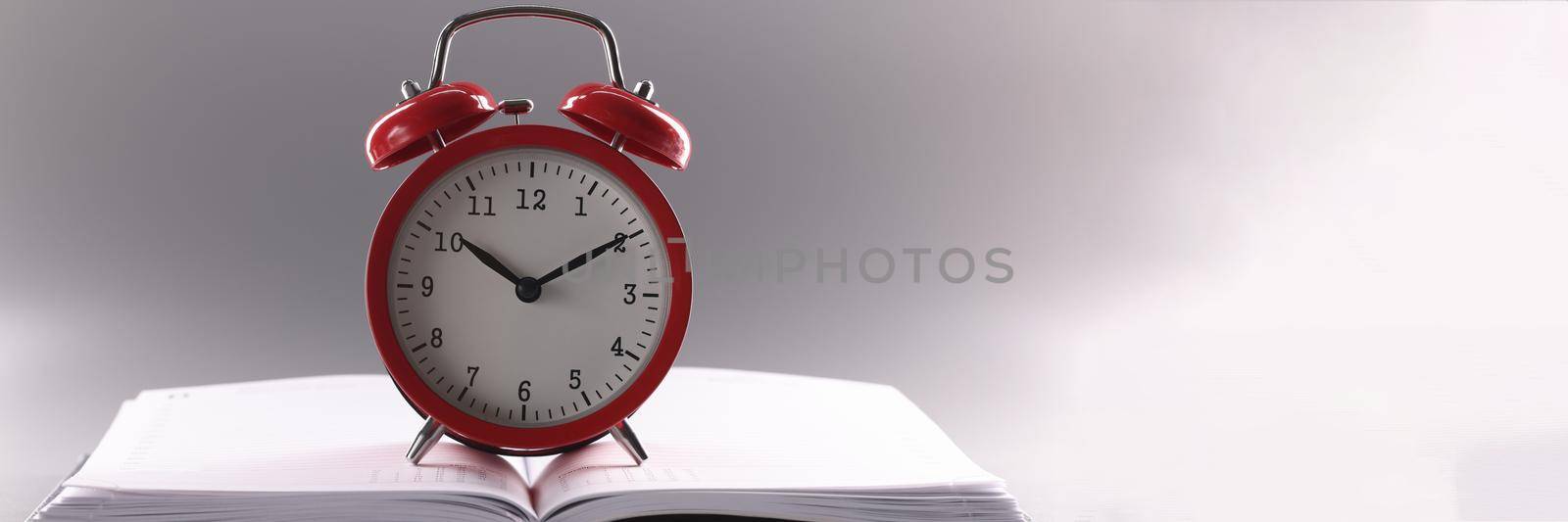 Close-up of red retro clock stand on top of open notebook or book. Time to learn idea, time to plan week on calendar. Time management, deadline concept