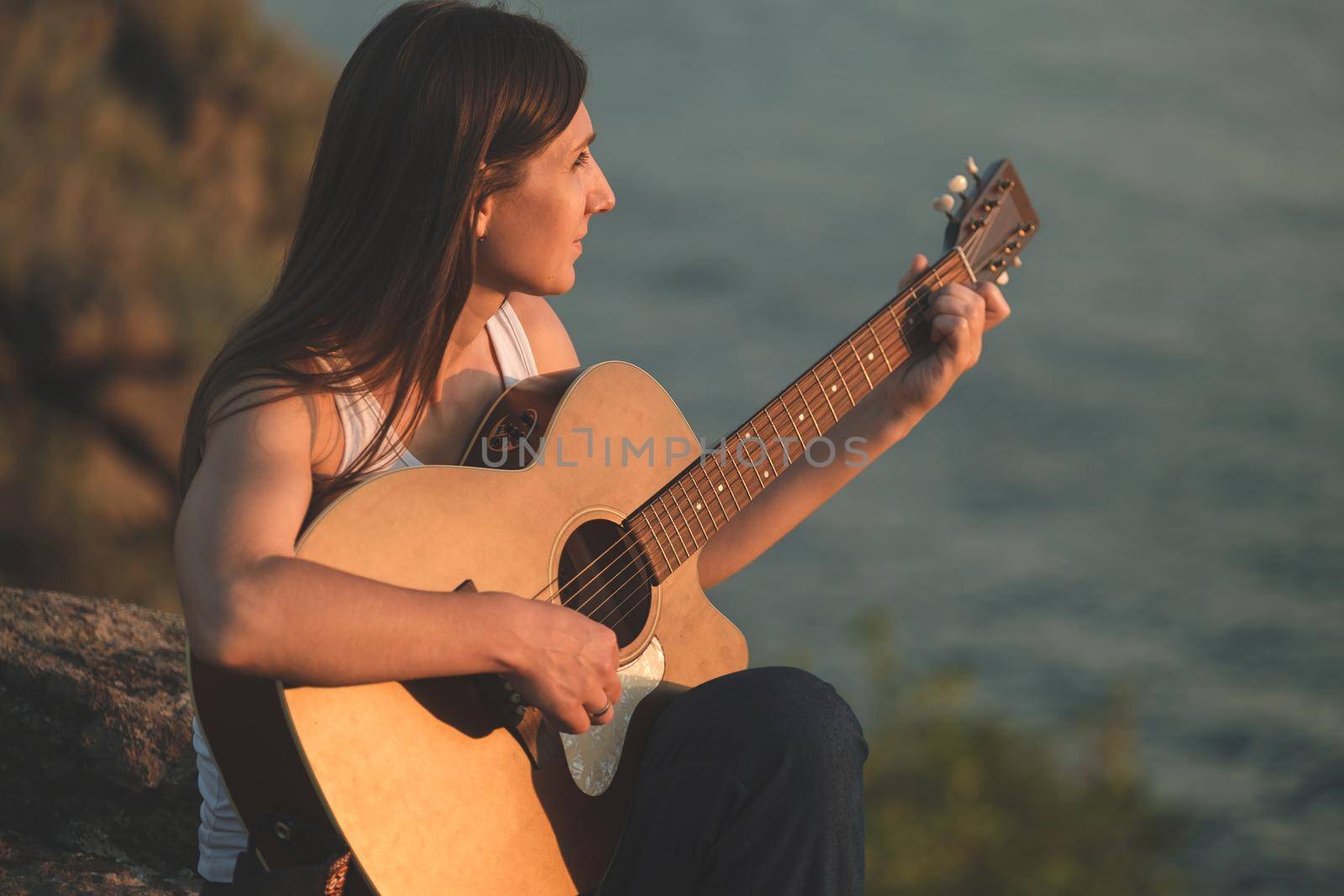A beautiful woman playing acoustic guitar sitting on the rock. Pretty girl practicing music on guitar. Female guitarist playing chords on acoustic guitar outdoor