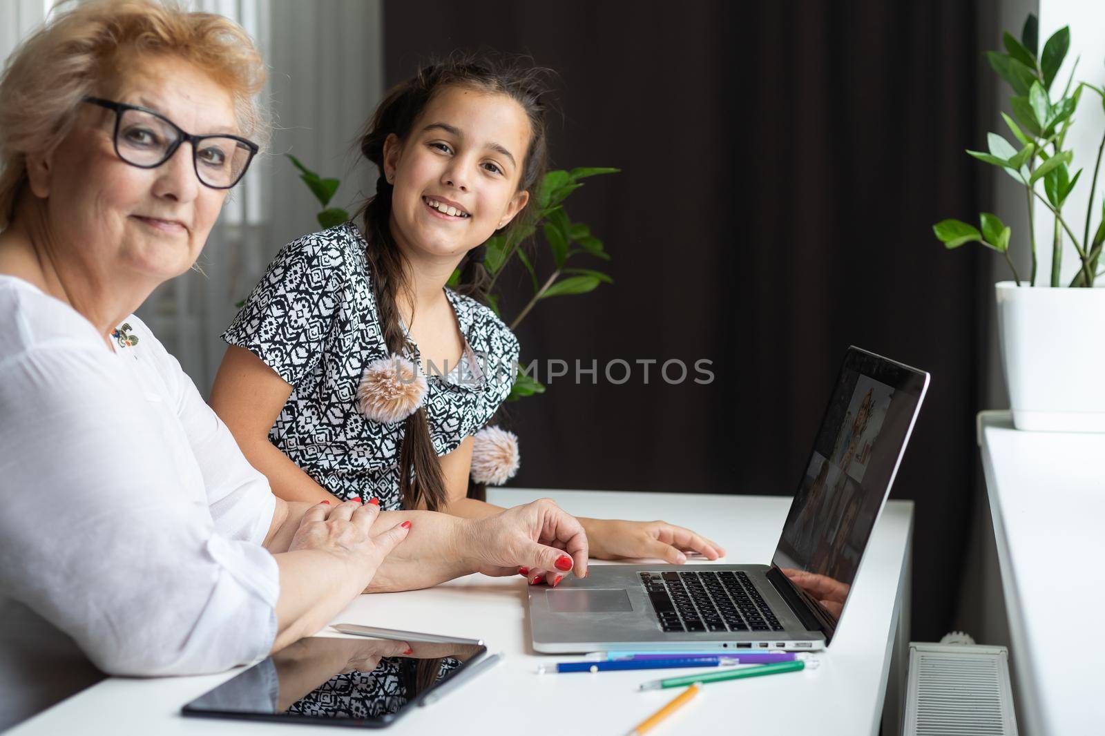 Portrait of happy grandmother and little granddaughter making video conference on pc sitting at table, waving hands at screen, greeting somebody, chatting with parents, enjoying online communication