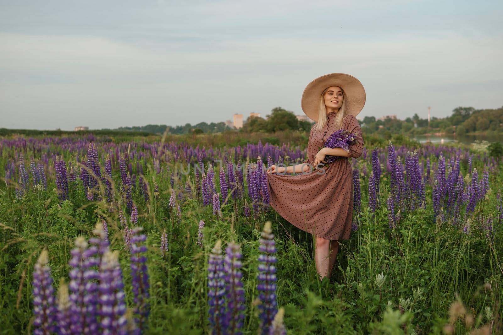 A beautiful woman in a straw hat walks in a field with purple flowers. A walk in nature in the lupin field.