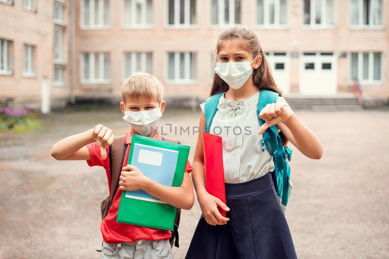 Unhappy schoolchildren, boy and girl in medical masks, stand near the school outdoors and showing thumbs down by VitaliiPetrushenko