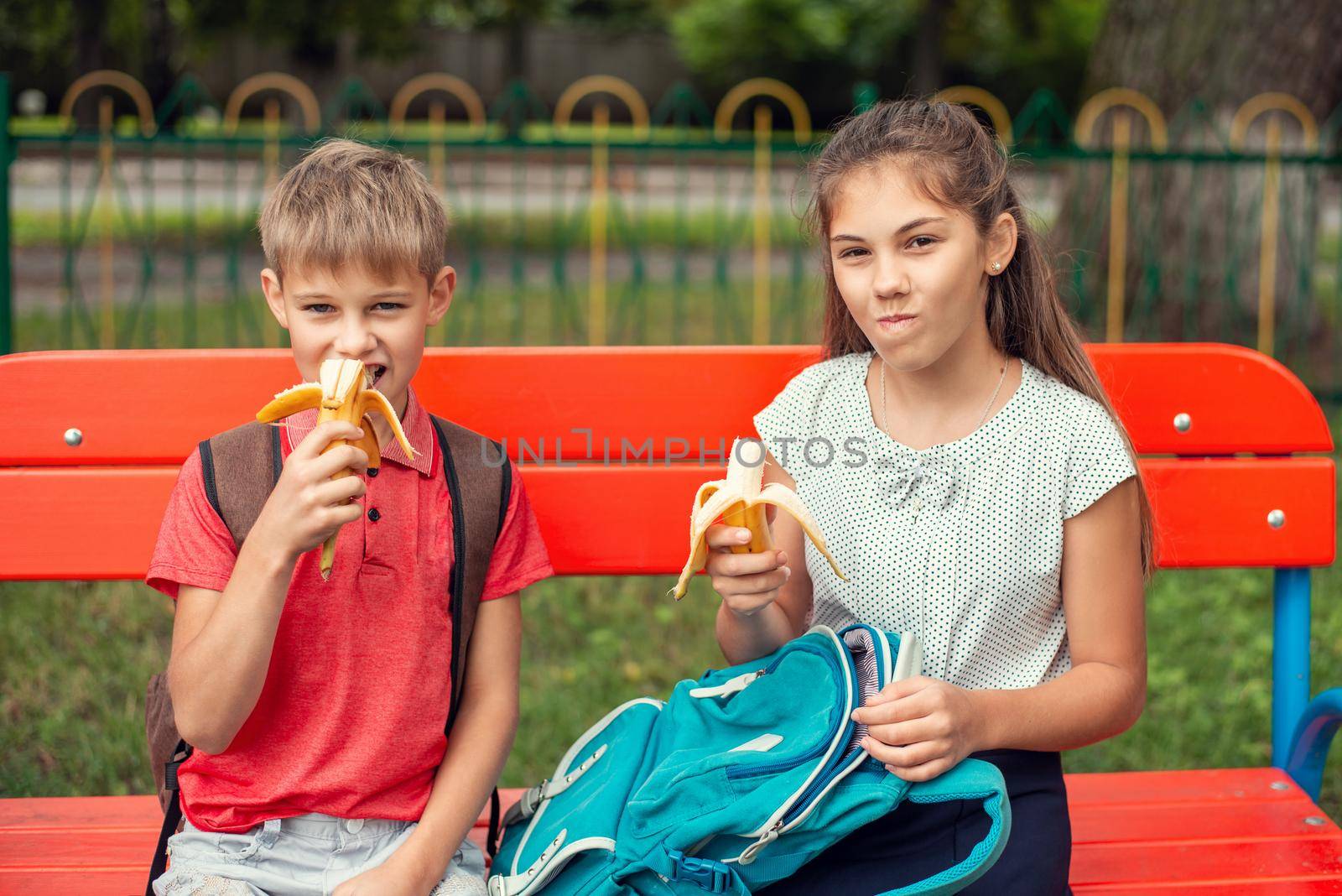 Two pupils friends, girl and boy, sitting on the bench near the school and eating bananas