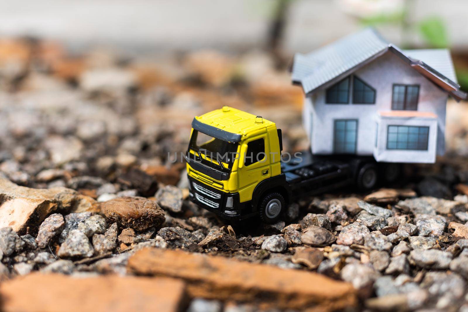 Close-up of a small toy truck on a tree stump carrying a miniature house model on top against a blurred forest background, front view. Countryside relocation service concept