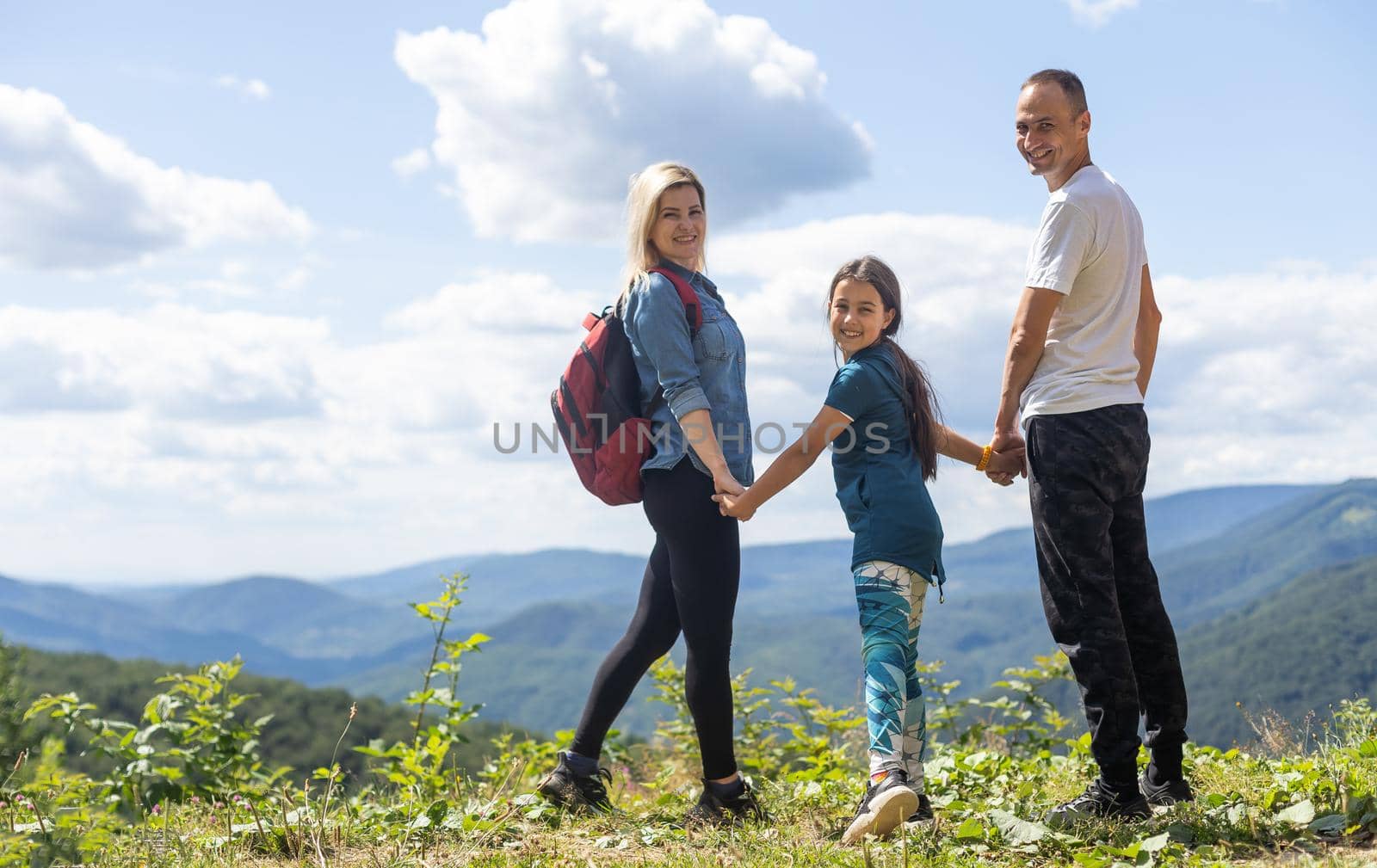 Family standing with arms around on top of mountain, looking at beautiful summer mountain landscape. People enjoying view