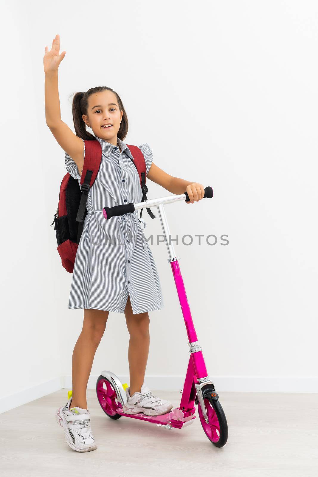 Full length profile shot of a schoolgirl with a backpack riding a scooter isolated on white background.