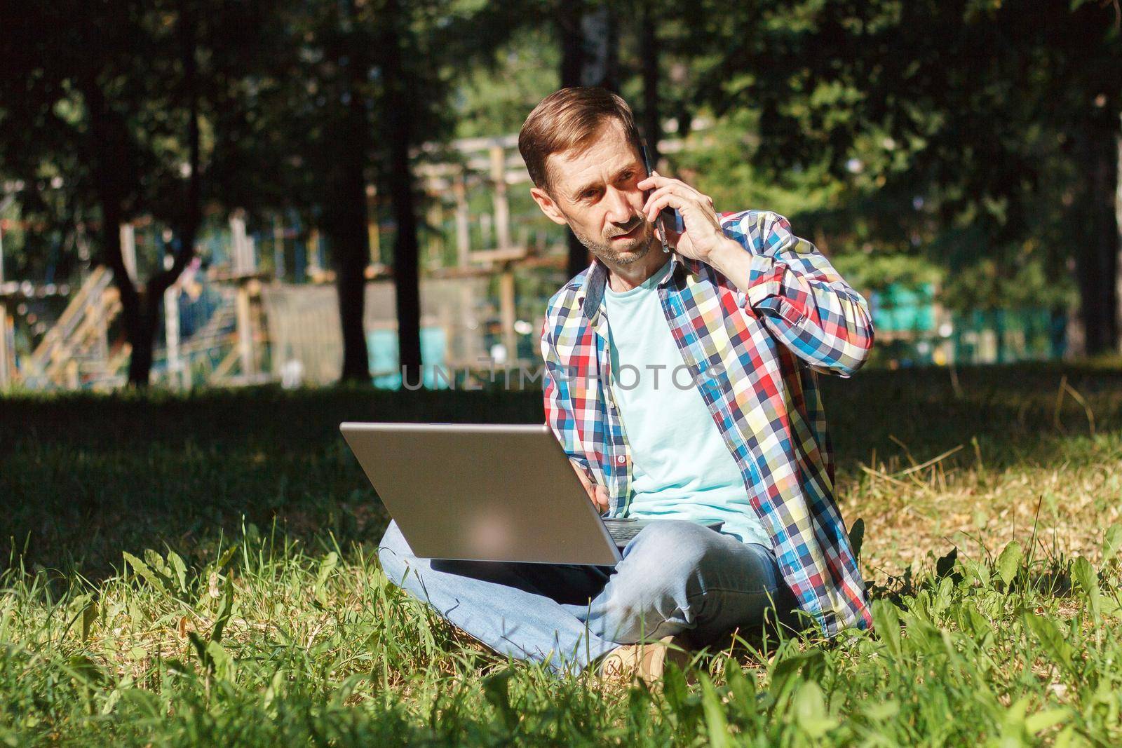 Adult man works on his computer in the park on the lawn.Remote work concept. The writer works remotely, enjoying nature.
