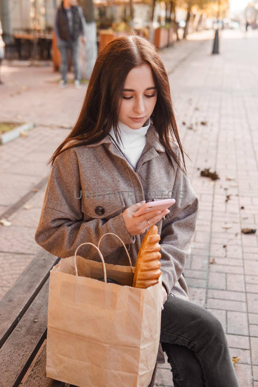 A stylish brunette woman walks around the autumn city. The brunette is sitting on a bench outside with a glass of coffee and a phone, holding a craft bag with a baguette.