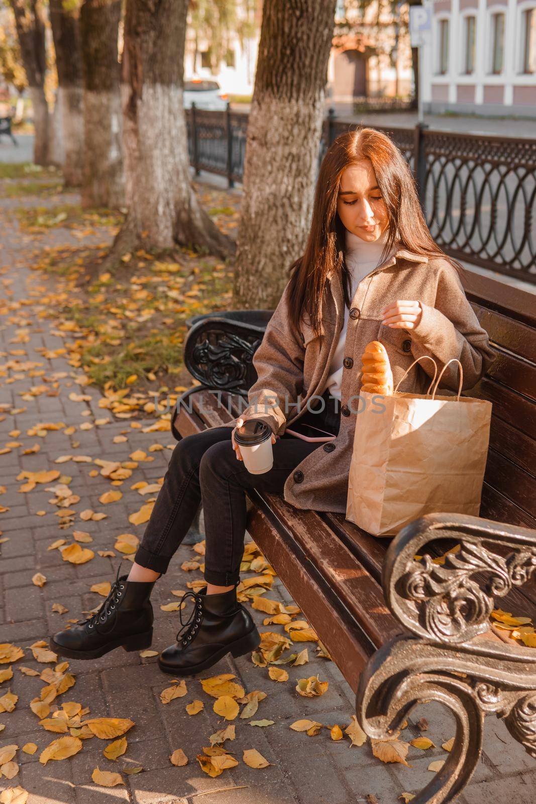 A stylish brunette woman walks around the autumn city. The brunette is sitting on a bench outside with a glass of coffee and a phone, holding a craft bag with a baguette.