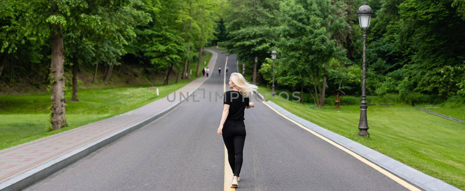 Joyful young lady in casual clothes walking along asphalt road in countryside, hitchhiking for ride outdoors. Lovely millennial woman traveling alone by autostop.