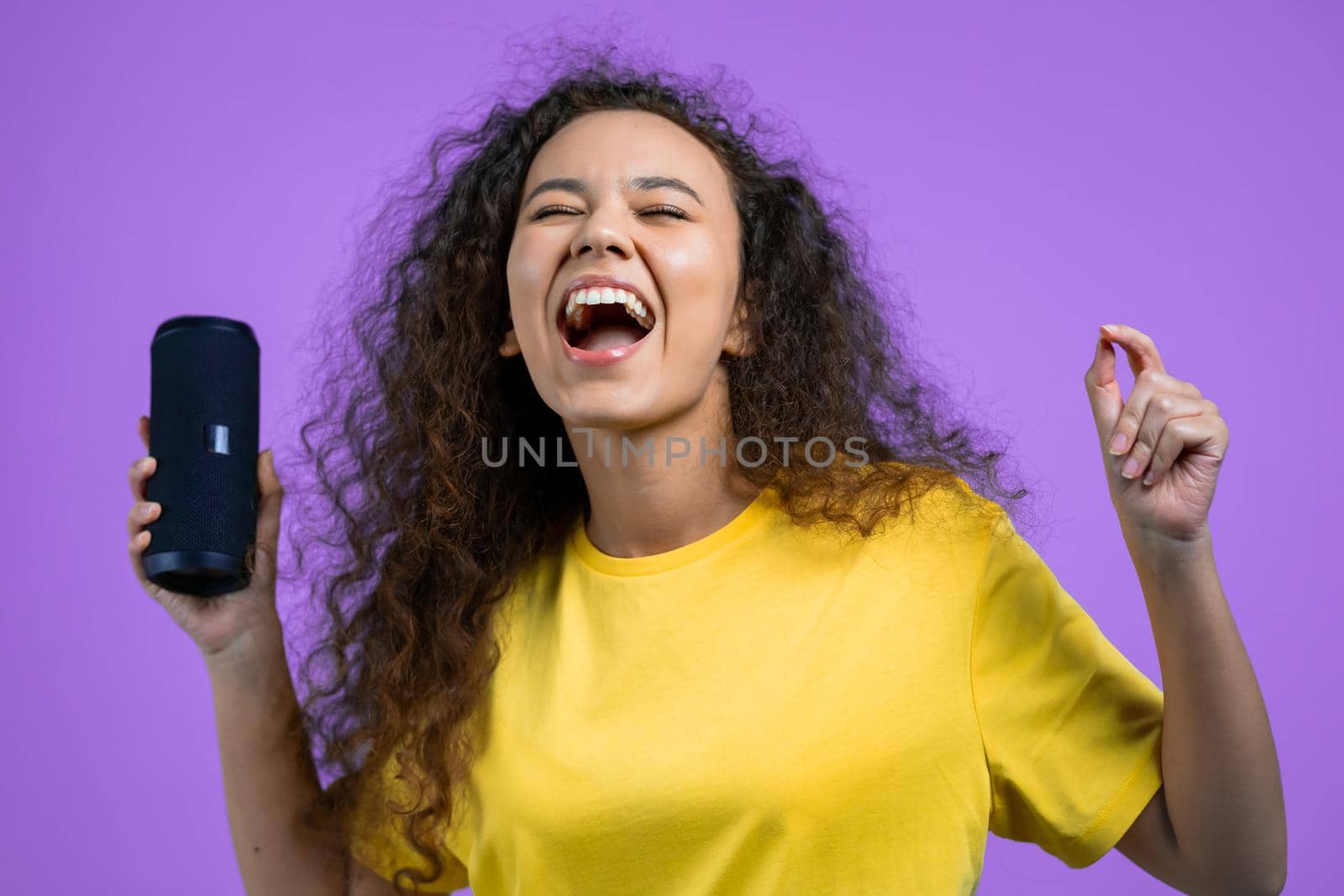 Mixed race woman listening to music by wireless portable speaker - modern sound system. Teenager dancing, enjoying on violet studio background. She moves to the rhythm of music. by kristina_kokhanova