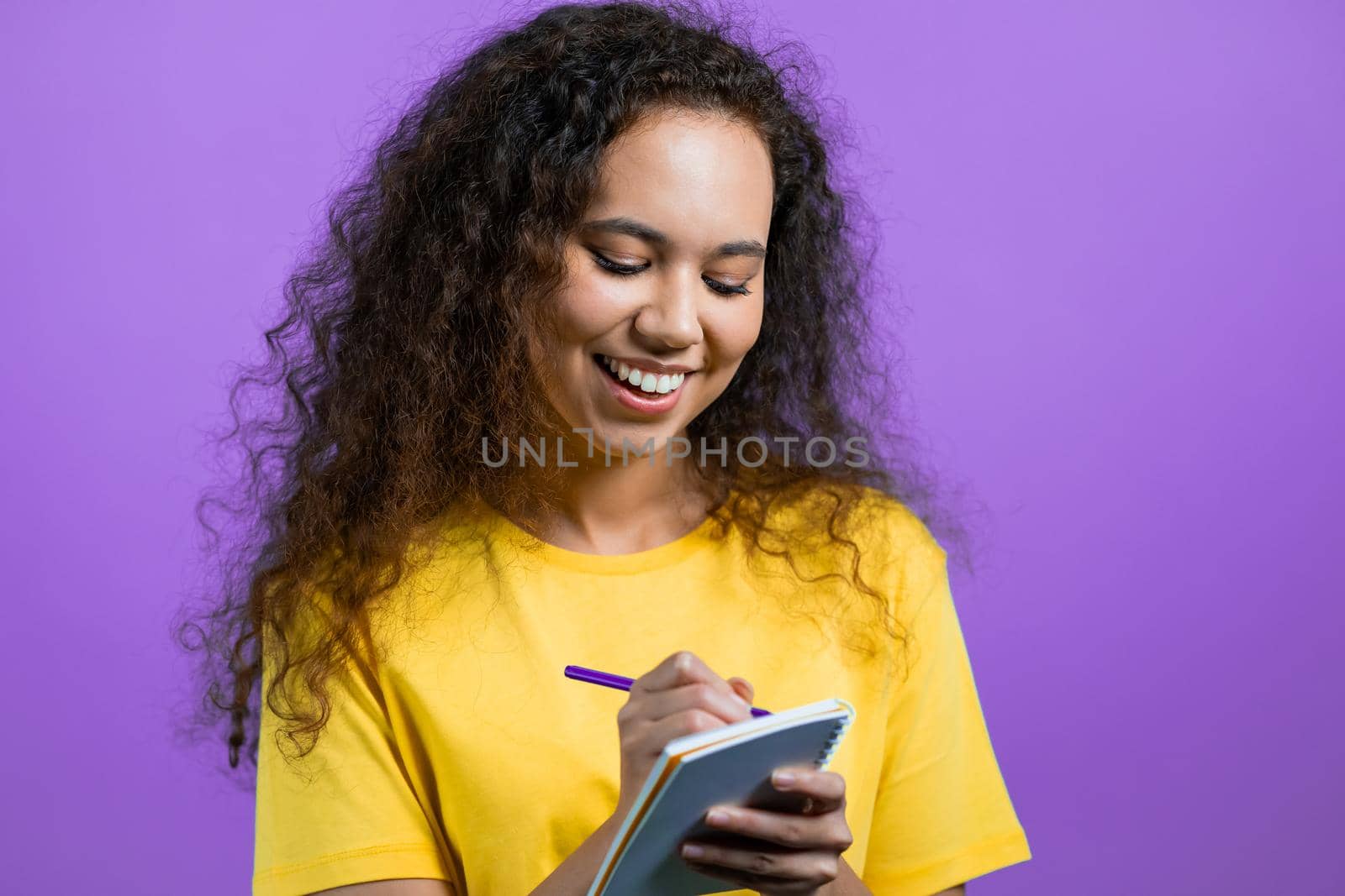 Mixed race woman making notes in planner, lady holding pen. She writes future plans and to-do list in notebook for week or month. Keeping personal diary on purple studio background. High quality photo