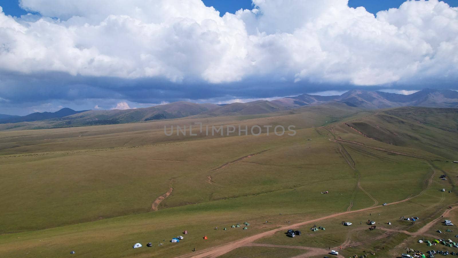 Big white clouds over green hills and mountains. Top view from the drone on endless fields. Roads are visible in places, herds of animals graze. A tent camp has been set up. Coniferous trees in gorge