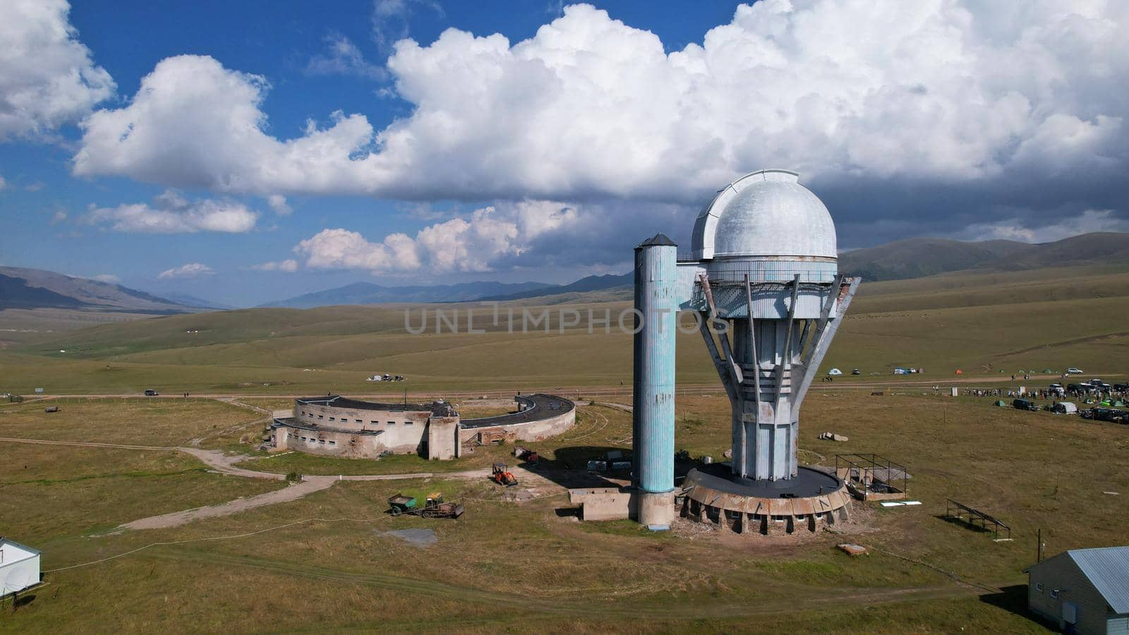The Assy-Turgen Observatory is high in the mountains. There is a tent camp next to the observatory. Large cumulus clouds in a blue sky. Yellow-green hills, forest in places. Top view from a drone