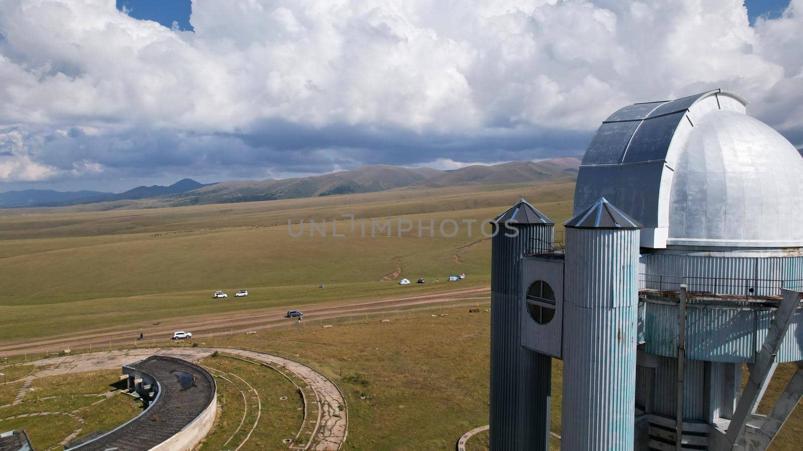 The Assy-Turgen Observatory is high in the mountains. There is a tent camp next to the observatory. Large cumulus clouds in a blue sky. Yellow-green hills, forest in places. Top view from a drone
