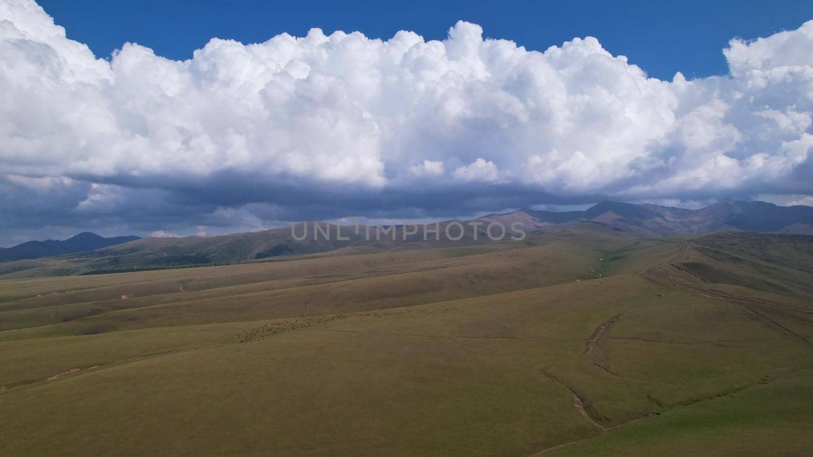 Big white clouds over green hills and mountains. Top view from the drone on endless fields. Roads are visible in places, herds of animals graze. A tent camp has been set up. Coniferous trees in gorge