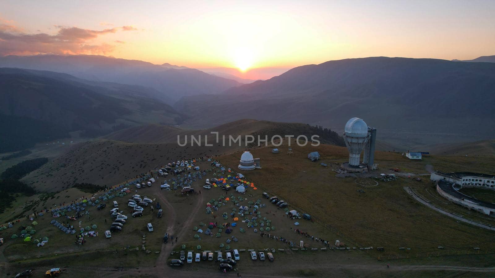 Two large telescope domes at sunset. Drone view of Assy-Turgen Observatory. Beautiful red sunset. Green hills and clouds. Tourists watch the sun. There is a large tent camp and cars nearby. Kazakhstan