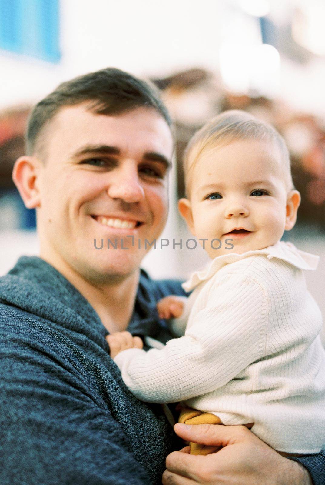 Dad with a baby in his arms stands near the house. Portrait by Nadtochiy